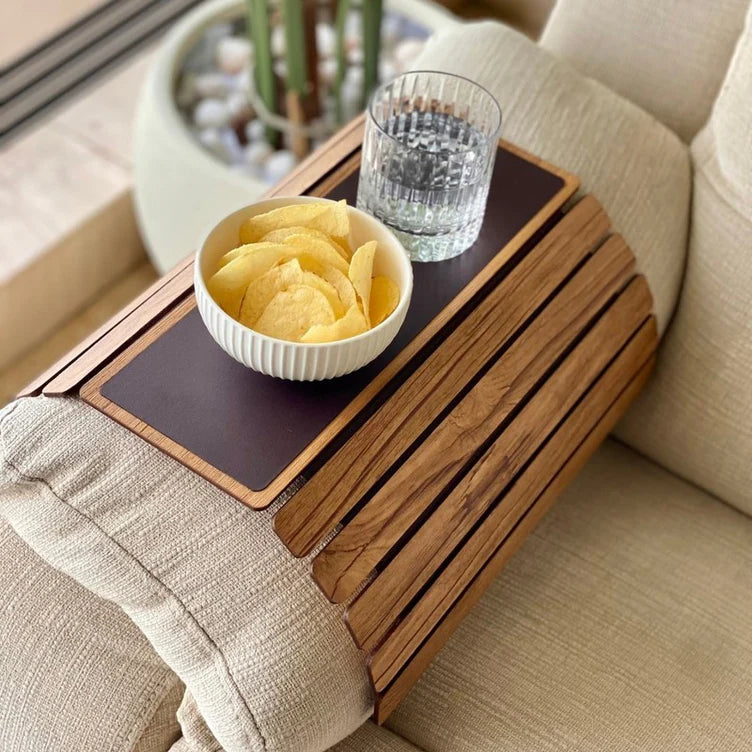 Wooden tray attached to a couch with a bowl of chips and a glass of water on a beige sofa.