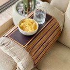 Wooden tray attached to a couch with a bowl of chips and a glass of water on a beige sofa.